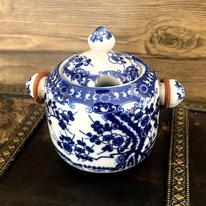 May include: A small, white ceramic sugar bowl with a blue and white floral design. The bowl has a lid with a small hole in the top and wooden handles.