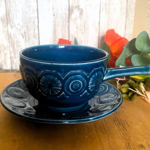 Puede incluir: Un tazón de sopa de cerámica azul verdoso con un platillo a juego. El tazón tiene una asa y ambas piezas presentan un patrón repetitivo de círculos y flores estilizadas.