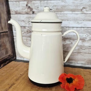 Può includere: Caffettiera in smalto color crema con base e finiture nere. La caffettiera vintage ha un beccuccio curvo e un manico. Un fiore arancione brillante con centro nero poggia sulla superficie di legno.