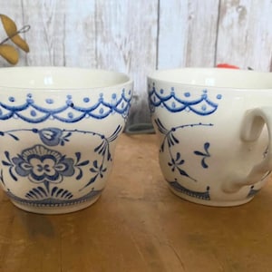 May include: Two white ceramic teacups with blue floral designs. The cups have a scalloped rim and a blue and white floral pattern. One cup has a handle, the other does not.