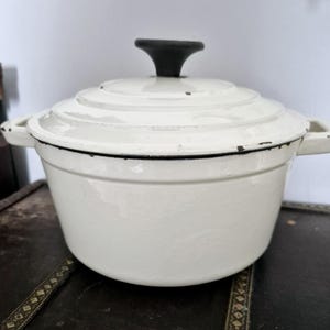 Puede incluir: Olla holandesa de hierro fundido color crema con un asa negra en la tapa. La olla tiene dos asas laterales y un borde negro. La olla esmaltada está diseñada para cocinar y hornear.