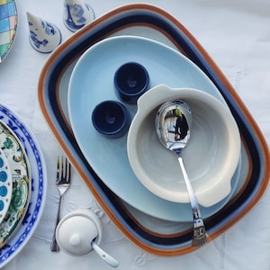 May include: A set of blue and white ceramic dishes with a brown and blue rimmed oval serving platter. The dishes are stacked on top of each other and include a small bowl, a large oval plate, and a small round plate. There are two small blue ceramic egg cups on the large oval plate. A silver spoon is in the small bowl.