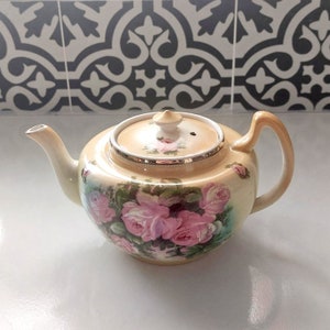Może przedstawiać: Kremowy czajnik z kwiatowym wzorem przedstawiającym różowe róże i zielone liście. Czajnik ma srebrną obwódkę wokół pokrywki i mały dzióbek.