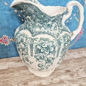 May include: A vintage white and teal ceramic pitcher with a floral pattern. The pitcher has a curved spout and a large, ornate handle. The floral design is repeated around the body of the pitcher.