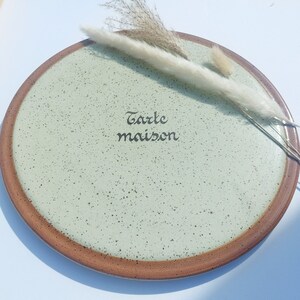 May include: A round ceramic plate with a speckled, pale green centre and a brown rim. The plate features the words "Tarte maison" in black script. Dried ornamental grass is placed on the plate.