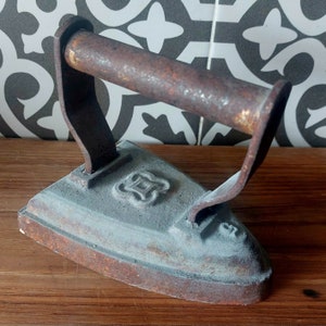 Puede incluir: Una plancha vintage de hierro fundido con un mango oxidado y un diseño decorativo en la base. La plancha tiene un aspecto desgastado y envejecido.