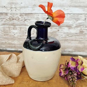 May include: A ceramic jug vase with a dark blue top and handle, and a cream-colored base. An orange flower is placed in the vase. The vase is on a wooden surface.