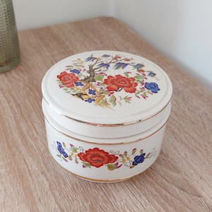 May include: A white ceramic jar with a lid, both decorated with a floral pattern in shades of red, blue, and green. The jar has a gold trim around the top and bottom.