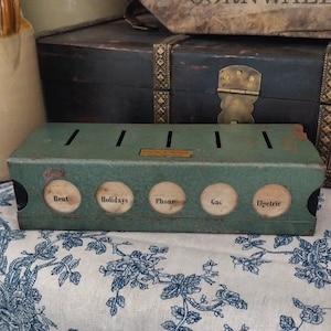 Könnte beinhalten: Vintage grüne Münzbank aus Metall mit Schlitzen und beschrifteten Abschnitten zum Sparen. Die Bank hat kreisförmige Öffnungen mit den Aufschriften "Rent", "Holidays", "Phone", "Gas" und "Electric". Ein kleines rechteckiges Etikett ist oben sichtbar.