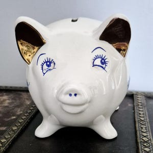 Può includere: Salvadanaio in ceramica bianca con occhi blu e ciglia, un muso dorato e orecchie dorate. Il salvadanaio ha una fessura nella parte superiore per le monete.