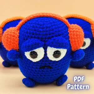 Op de afbeelding: Drie blauwe gehaakte amigurumi-speelgoedjes met oranje gehaakte oorwarmers. De speelgoedjes hebben witte ogen en een fronsende uitdrukking. De speelgoedjes staan op een lichtoranje achtergrond. PDF-patroon.