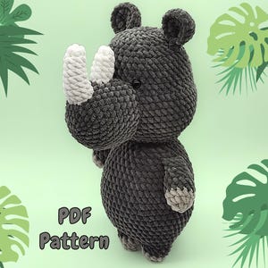 Op de afbeelding: Een gehaakt grijs neushoorn amigurumi speelgoed met witte hoorns. Het speelgoed zit op een groene achtergrond met groene bladeren.