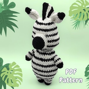 May include: A crocheted zebra amigurumi toy with black and white stripes. The toy has a black nose and black eyes. The background is a light green with green leaves.