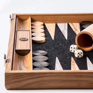 May include: A wooden backgammon set with a dark playing surface and light-colored wooden game pieces. Two dice and a wooden dice cup are also visible. The set is made of light brown wood with a dark brown playing surface.
