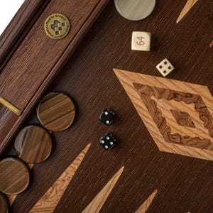 Könnte beinhalten: Ein Backgammon-Brett aus Holz mit brauner und beigefarbener Oberfläche. Das Brett hat eine rautenförmige Einlage in der Mitte. Auf dem Brett befinden sich schwarze und weiße Würfel sowie braune Spielsteine. Das Brett hat eine Messingplakette mit der Aufschrift "Handgefertigt in Griechenland seit 2015".