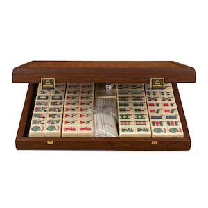 Pode incluir: Um conjunto de Mahjong de madeira numa caixa retangular castanha escura com tampa articulada. O interior contém filas de peças de cor branco-sujo com símbolos gravados a vermelho, verde e preto, e um saco com varetas de contagem brancas.