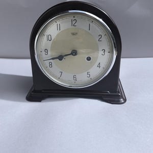 Puede incluir: Un reloj vintage marrón con una esfera color crema y manecillas negras. El reloj tiene un bisel plateado y una base rectangular. La esfera muestra los números del 1 al 12. Es un modelo Smiths Enfield.