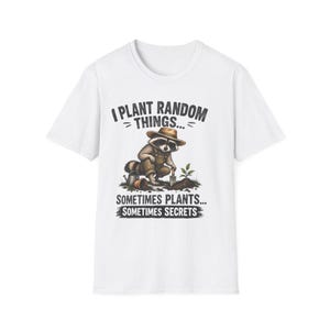 May include: A white short-sleeve t-shirt featuring a graphic of a raccoon wearing a brown hat and overalls, digging in soil with a trowel. The text reads: I PLANT RANDOM THINGS... SOMETIMES PLANTS... SOMETIMES SECRETS in a dark grey font.