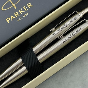 May include: A set of two silver-toned metal Parker pens housed in a dark blue and cream-colored presentation box. One pen is engraved with the name Dr. Harold Greene, and the other is engraved with the name Harold Greene. The box features the gold Parker logo.