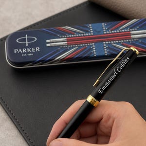May include: A black ballpoint pen with gold-colored accents and the name Emmanuel Collins engraved on the barrel. The pen is positioned in front of a rectangular metal Parker brand storage case featuring a Union Jack flag design.