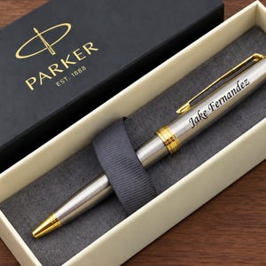 May include: A silver Parker ballpoint pen with gold-colored accents and a clip, featuring the engraved name Jake Fernandez on the barrel. The pen is presented in a cream-colored box with a dark gray interior and a black Parker gift box lid.