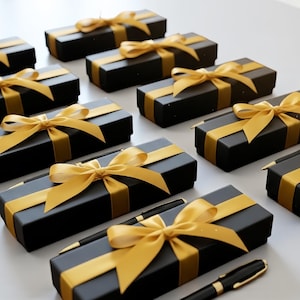 May include: Several black gift boxes with gold ribbon bows are arranged on a white surface. The boxes are rectangular and have a gold ribbon wrapped around them. Some boxes have a black pen with gold accents placed on top.