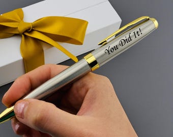 May include: A silver-toned ballpoint pen with gold-colored accents and a clip, featuring the engraved text "You Did It!" on the barrel. The pen is presented alongside a white rectangular gift box adorned with a decorative gold ribbon bow.
