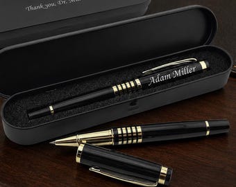 May include: A black and gold ballpoint pen with the name Adam Miller engraved on the barrel, resting inside a black case. The case lid is engraved with the text Thank you, Dr. Miller. A second matching pen and its cap are placed in front.