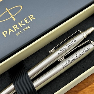 May include: Two silver Parker pens in a gift box. One pen is engraved with "Lucy Smith," and the other with "Published Author." The box is navy blue with the Parker logo in gold and the words "EST. 1888."