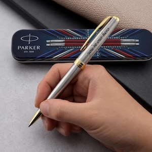 May include: A silver-toned Parker ballpoint pen with gold-colored accents and the engraved text "you're our hero" on the barrel. The pen is positioned in front of a rectangular metal case featuring a blue, white, and red graphic design.