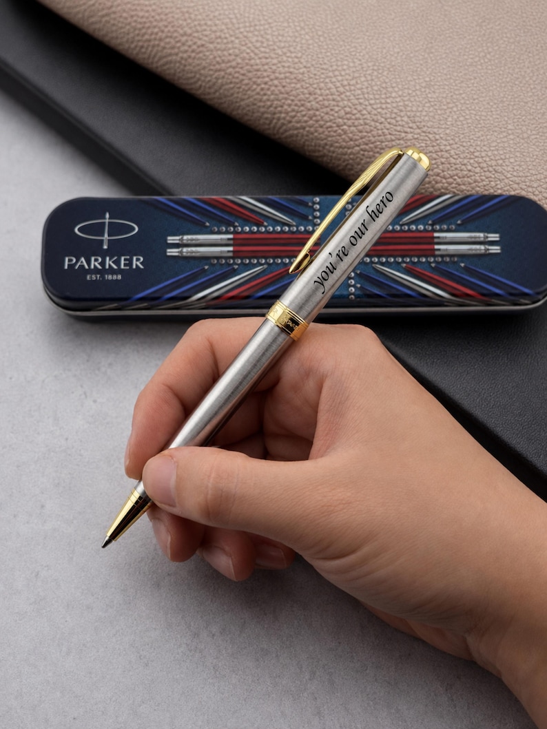 May include: A silver-toned Parker ballpoint pen with gold-colored accents and the engraved text "you're our hero" on the barrel. The pen is positioned in front of a rectangular metal case featuring a blue, white, and red graphic design.