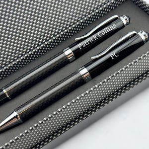 May include: A set of two black pens with silver accents, presented in a textured gray box. One pen is engraved with "Patrick Collins," and the other with "PC." The pens have a carbon fiber pattern and are designed for writing.