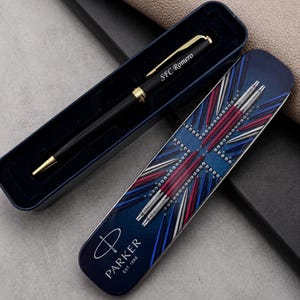 May include: A black ballpoint pen with gold-tone accents and the engraved text SFC Romero, resting in a dark blue case. The lid features a graphic of multiple pens arranged in a starburst pattern, the Parker logo, and the text PARKER EST. 1888.