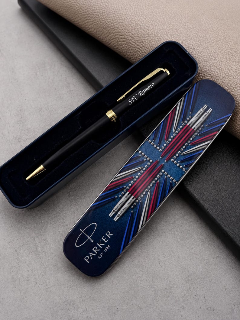 May include: A black ballpoint pen with gold-tone accents and the engraved text SFC Romero, resting in a dark blue case. The lid features a graphic of multiple pens arranged in a starburst pattern, the Parker logo, and the text PARKER EST. 1888.