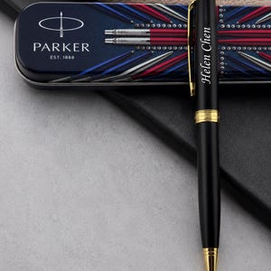 May include: A black ballpoint pen with gold-colored trim and the name Helen Chen engraved on the barrel. The pen is positioned in front of a rectangular metal storage tin featuring the Parker logo, the text EST. 1888, and a red, white, and blue design.