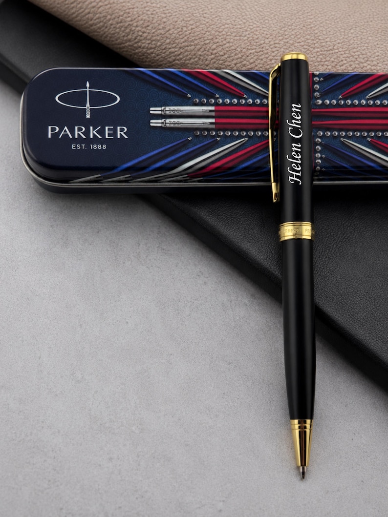 May include: A black ballpoint pen with gold-colored trim and the name Helen Chen engraved on the barrel. The pen is positioned in front of a rectangular metal storage tin featuring the Parker logo, the text EST. 1888, and a red, white, and blue design.