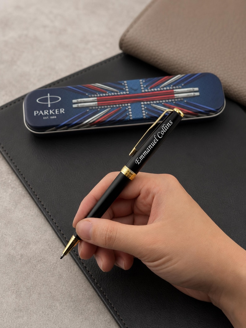 May include: A black ballpoint pen with gold-colored accents and the name Emmanuel Collins engraved on the barrel. The pen is positioned in front of a rectangular metal Parker brand storage case featuring a Union Jack flag design.