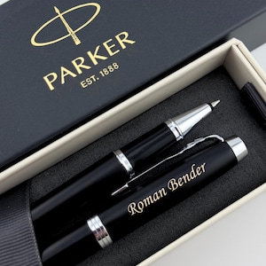 May include: A black Parker rollerball pen with silver-tone accents and a pocket clip, personalized with the name Roman Bender engraved in gold. The pen is presented in a black gift box with the gold Parker logo and the text EST. 1888 printed on top.