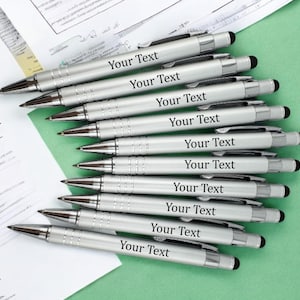 May include: A collection of silver pens with black ink tips and touch screen stylus tips. Each pen is inscribed with the words "Your Text" in black lettering. The pens are arranged on a green surface with documents in the background.