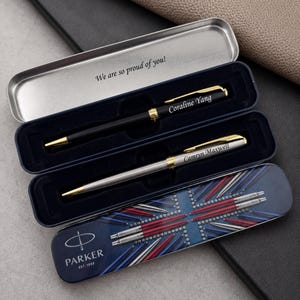May include: Two Parker pens in a metal case. One pen is matte black with gold accents and the name Coraline Yang. The other is silver with gold accents and the name Camryn Maxwell. The case lid features a Union Jack design and the text We are so proud of you!