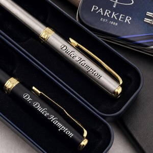 May include: A set of two pens in a dark blue case. One pen is brushed silver with gold accents and engraved with Dulce Hampton. The other is matte black with gold accents and engraved with Dr. Dulce Hampton. A Parker brand tin is visible nearby.
