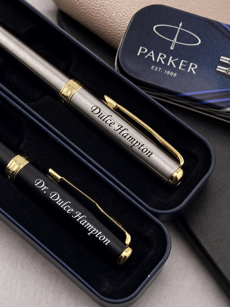 May include: A set of two pens in a dark blue case. One pen is brushed silver with gold accents and engraved with Dulce Hampton. The other is matte black with gold accents and engraved with Dr. Dulce Hampton. A Parker brand tin is visible nearby.