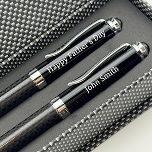May include: Two black pens with silver accents are displayed in a gift box. One pen reads "Happy Father's Day" and the other reads "John Smith" in white lettering. The pens have a carbon fiber pattern.