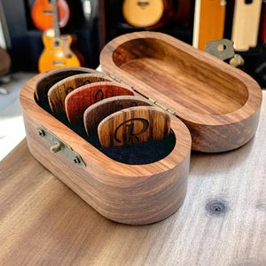 May include: A wooden box with a hinged lid, containing a set of six guitar picks. The picks are made of wood and have different colors and patterns. The box is made of dark wood and has a brass latch.