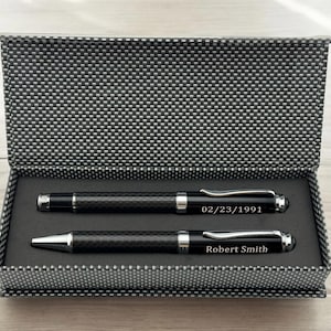 May include: A black gift box with a gray carbon fiber pattern on the outside. The box is open and contains two black pens with silver accents. The top pen has the date "02/23/1991" engraved on it. The bottom pen has the name "Robert Smith" engraved on it.
