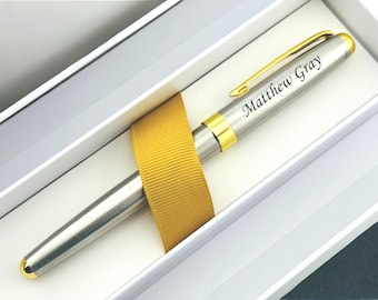May include: A silver-toned metal pen with gold-colored accents and a gold clip, featuring the engraved name Matthew Gray on the cap. The pen is secured with a mustard yellow ribbon inside a white rectangular presentation box.
