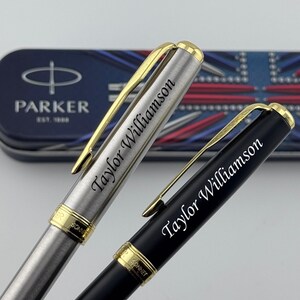 May include: Two Parker Sonnet pens with gold-tone clips and trim. One pen has a brushed stainless steel finish, and the other has a matte black finish. Both pens are engraved with the name Taylor Williamson and rest before a Parker branded tin.