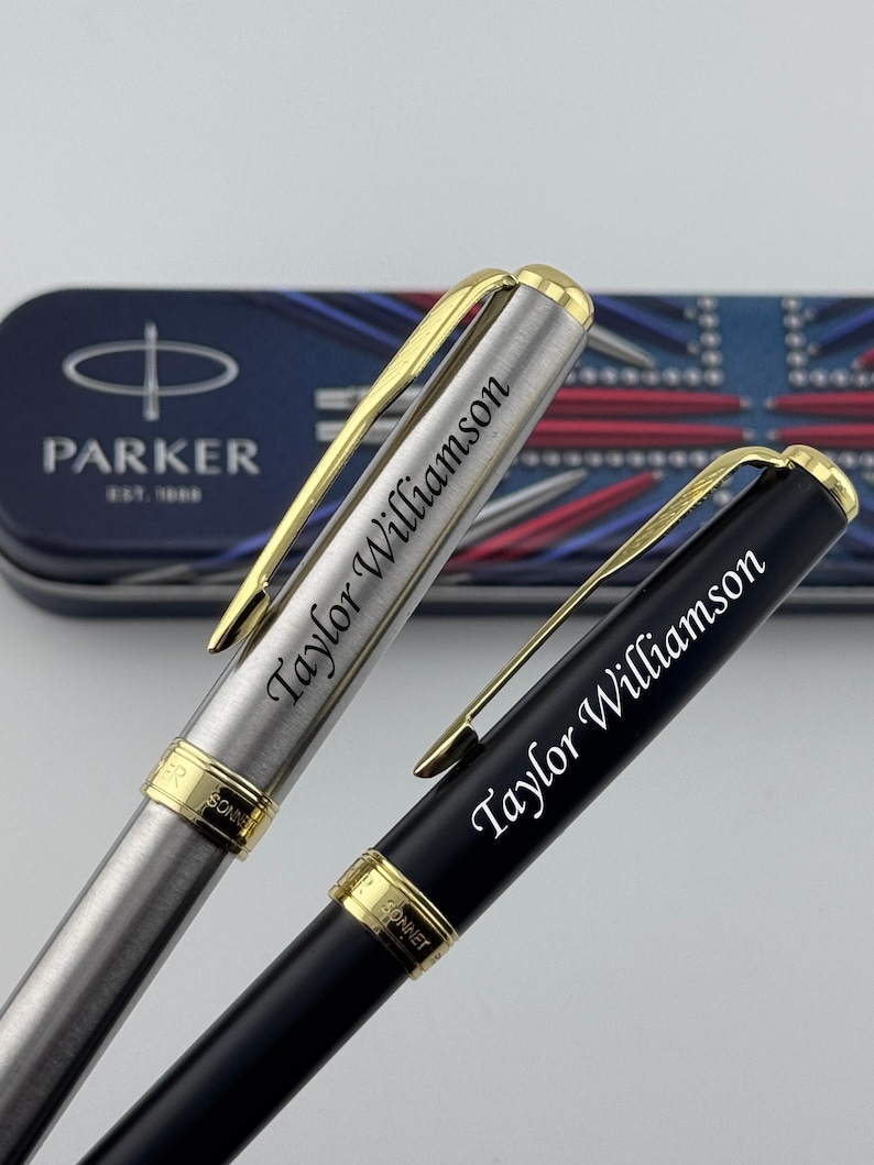 May include: Two Parker Sonnet pens with gold-tone clips and trim. One pen has a brushed stainless steel finish, and the other has a matte black finish. Both pens are engraved with the name Taylor Williamson and rest before a Parker branded tin.