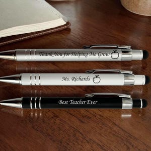 May include: Three pens with stylus tips are displayed on a wooden surface. Two pens are silver with black text and an apple graphic. The third pen is black with silver accents and the text "Best Teacher Ever".