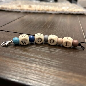 May include: A beaded keychain spelling out the word "WOOL". The keychain features wooden cube beads with letters, interspersed with round beads in various colors including blue, gray, and brown. A silver-colored clasp is attached.